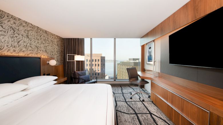 A modern hotel room with a large bed, a wall-mounted TV, a desk with a chair, and a window offering a view of city buildings and water.