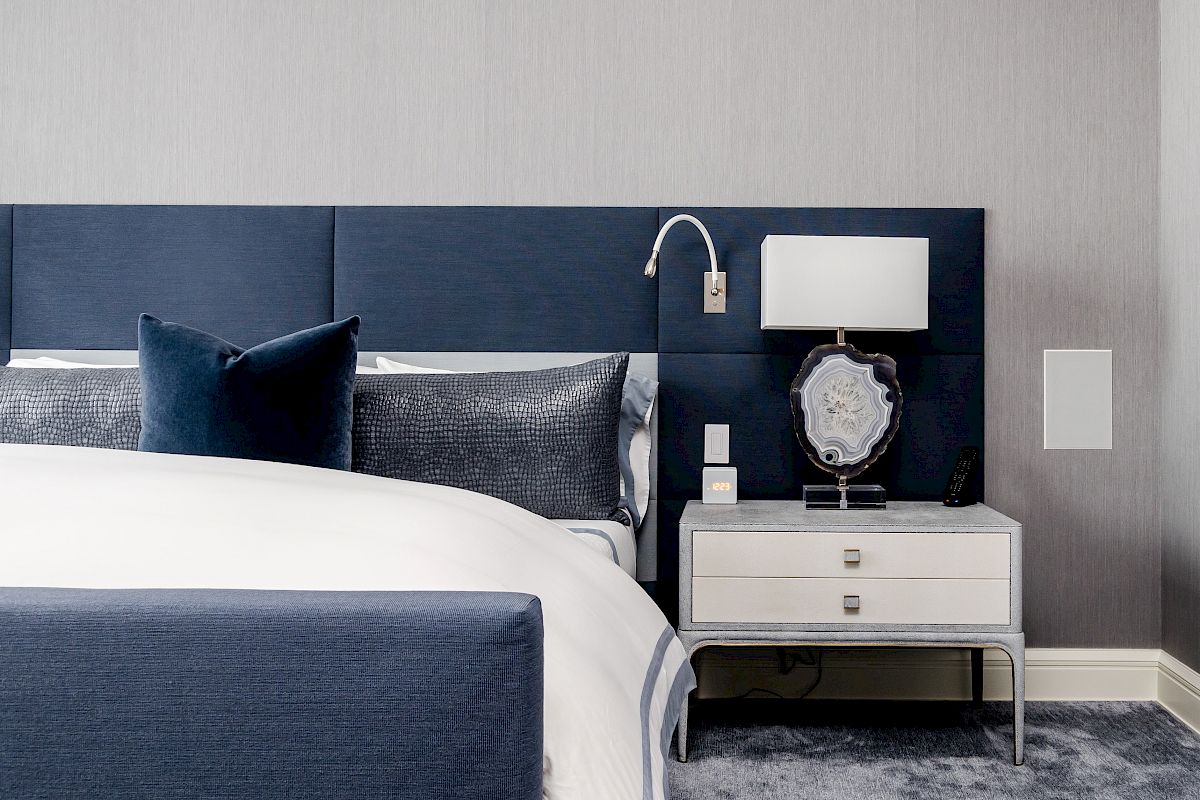 A modern bedroom with a blue and white theme, featuring a bed, a blue headboard, and a nightstand with a lamp and decorative items.