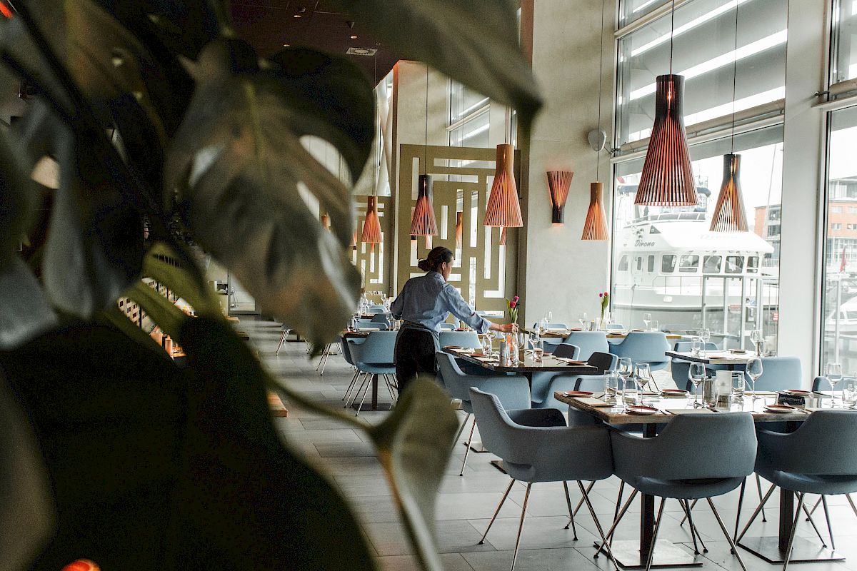 A person prepares tables in a modern, well-lit restaurant with stylish decor and hanging lights, as seen through some indoor plants.